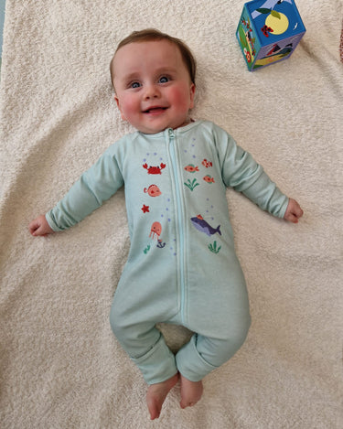 Bébé allongé sur tapis et souriant portant le pyjama "nager au lieu des coraux" de la collection mer.